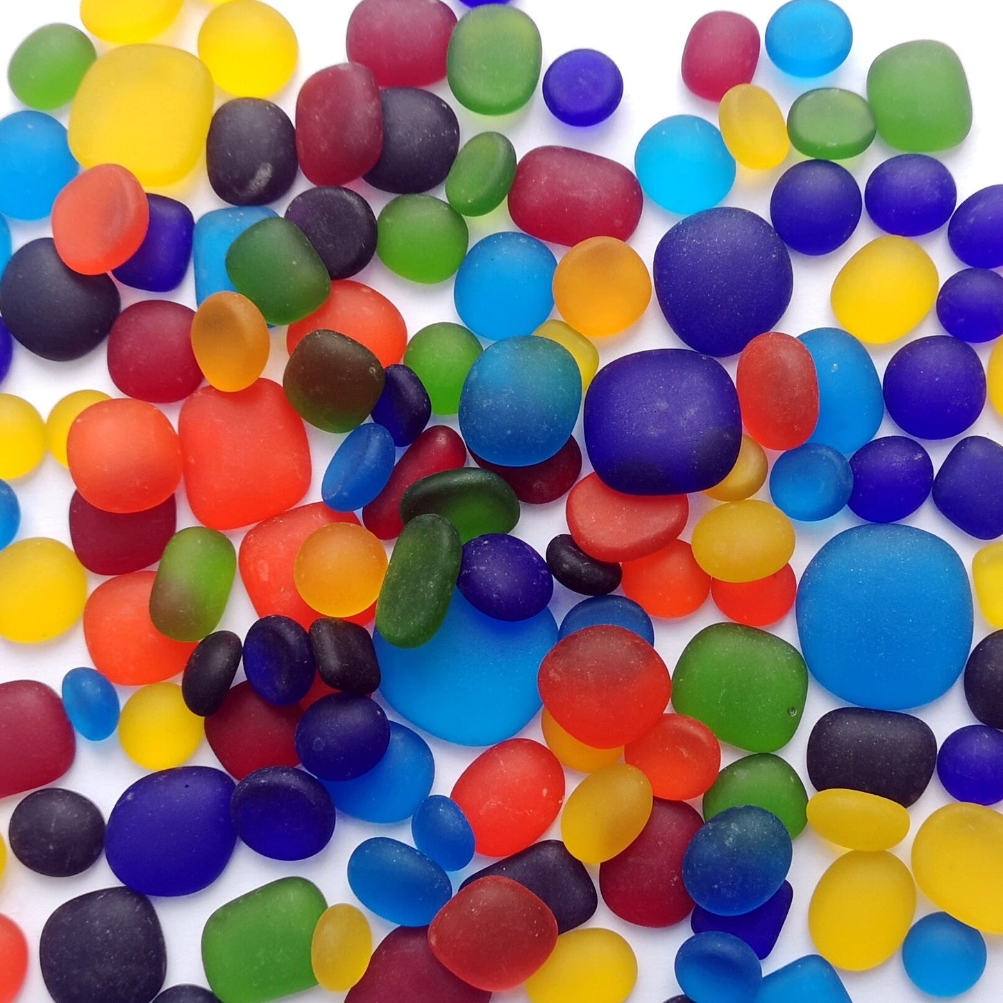 Vibrant, rainbow-coloured tumbled glass pieces on a white background, focusing on their raw, frosted texture for eclectic craft projects