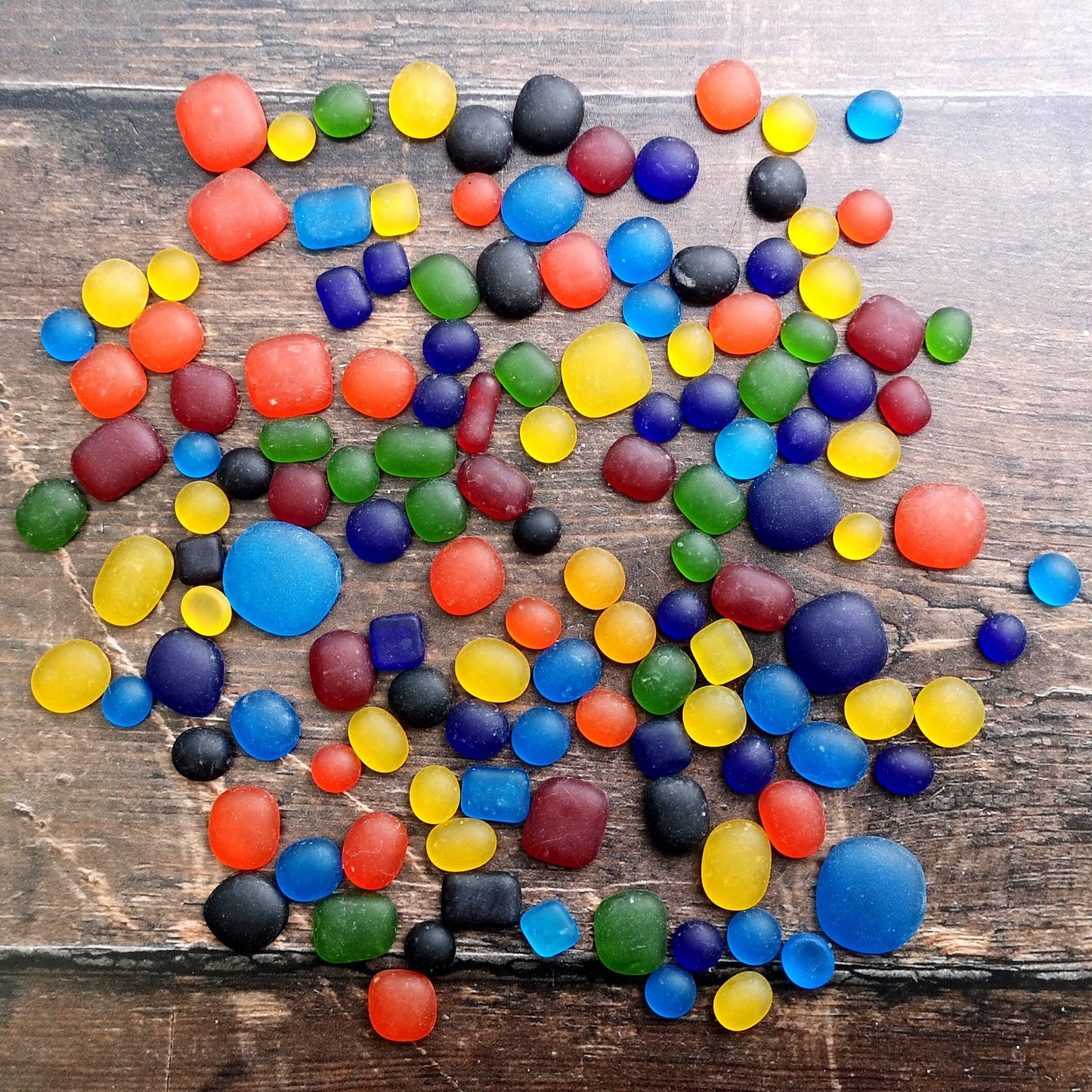 Multicolored tumbled glass in a rainbow palette, laid out on a dark surface, showcasing their frosty, sea glass-like finish for creative uses
