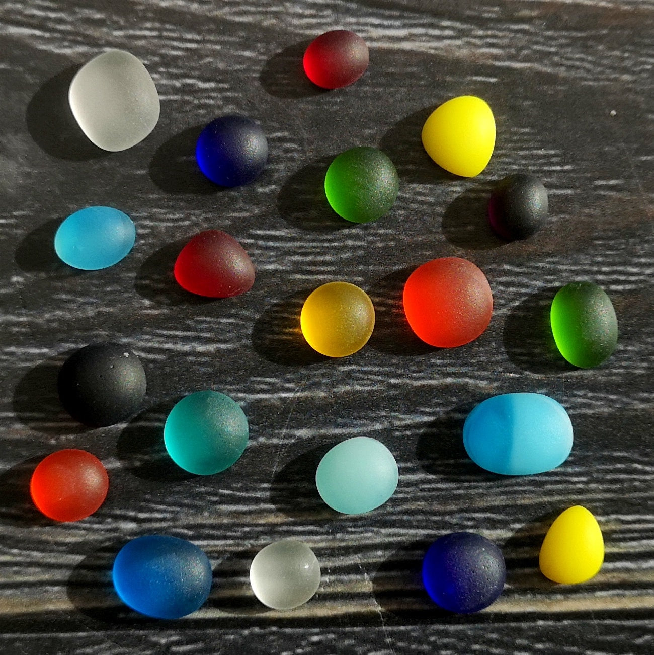 Tiny tumbled glass pieces in a spectrum of colors, arranged on a dark surface, showcasing their frosty, weathered appearance for unique crafts