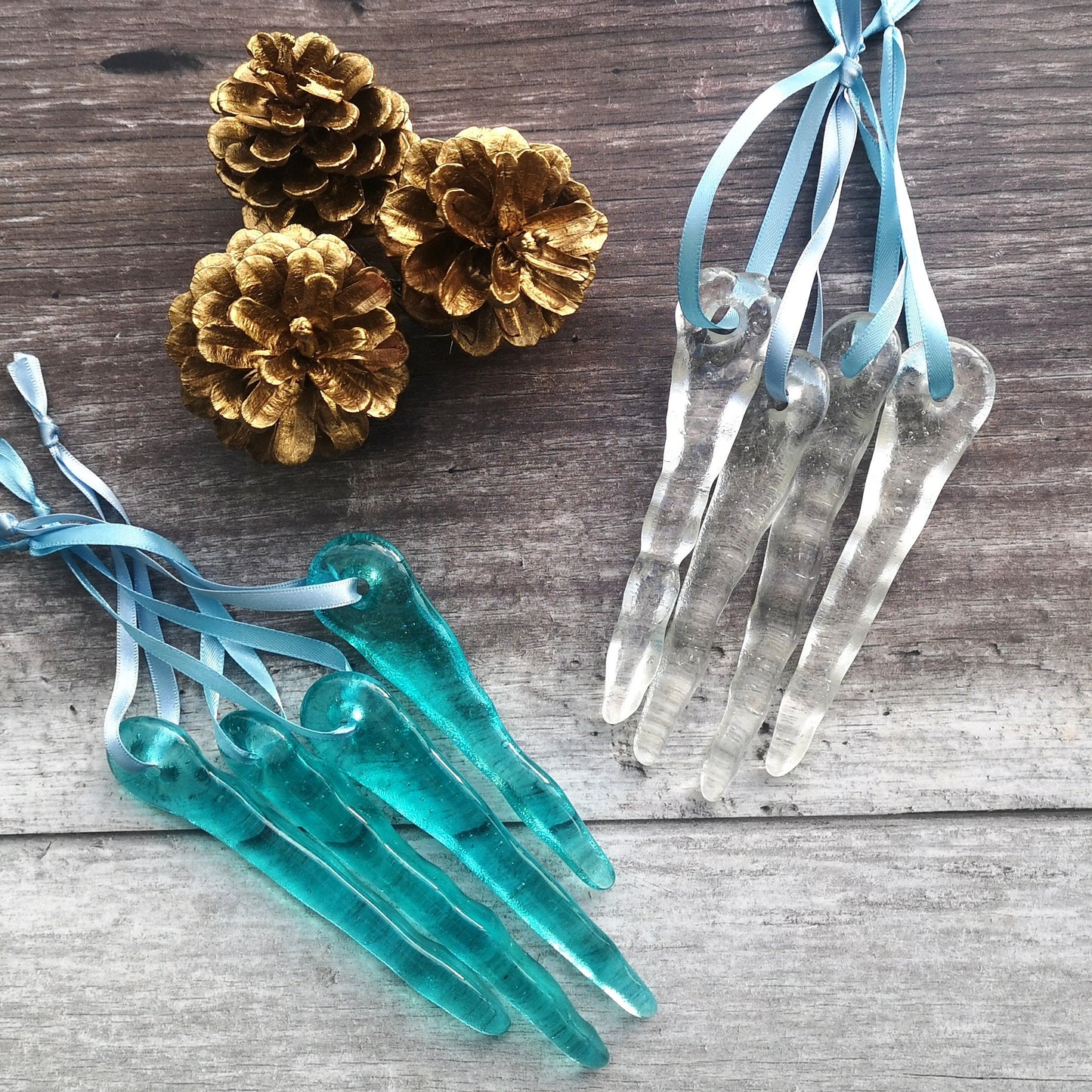 Two sets of four glass icicles in aqua blue and clear, arranged on a rustic wooden surface, highlighting their slender shape and sparkle.