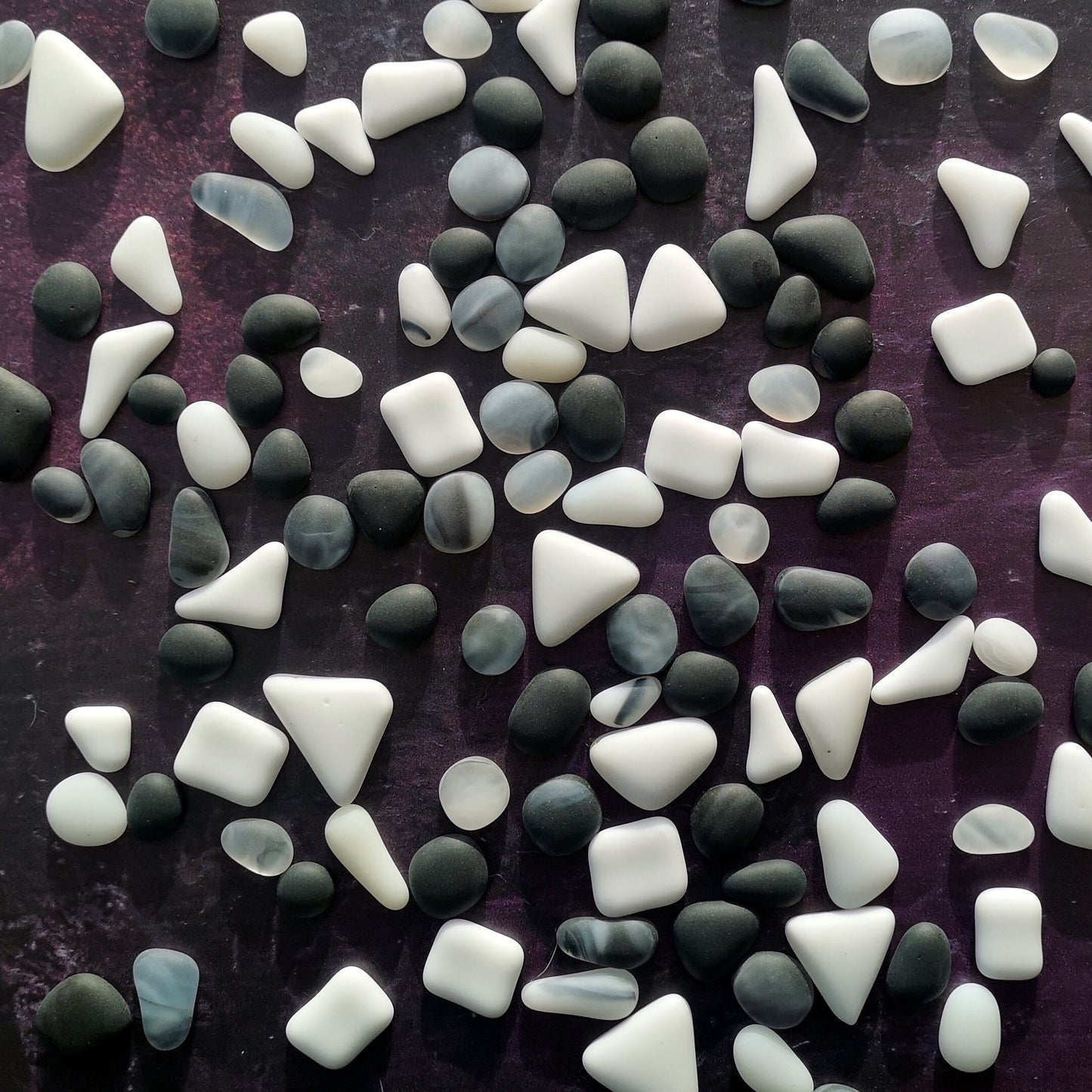 White and Balck tumbled glass assortment with a smooth finish, spread on a dark surface, focusing on their raw texture ideal for artistic designs.