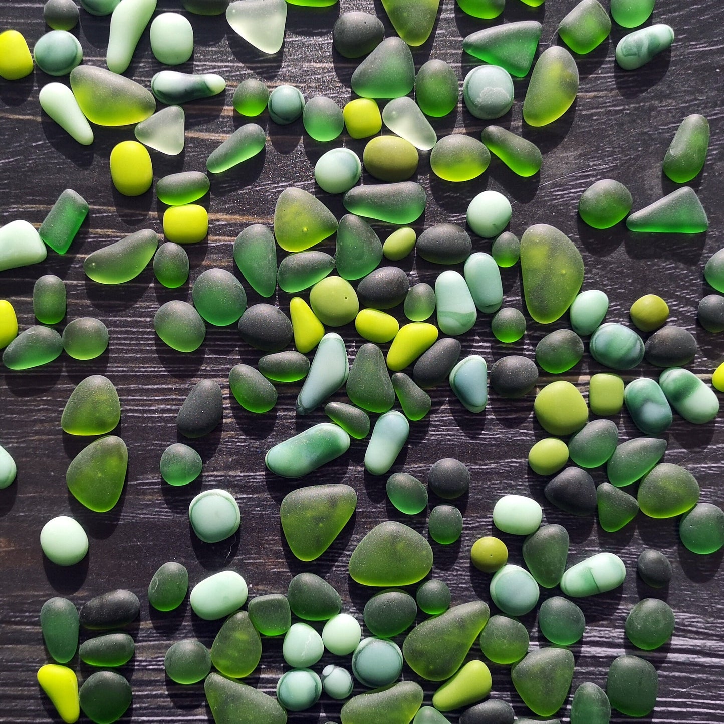 Multicolored tumbled glass in a rgreen palette, laid out on a dark surface, showcasing their frosty, sea glass-like finish for creative uses