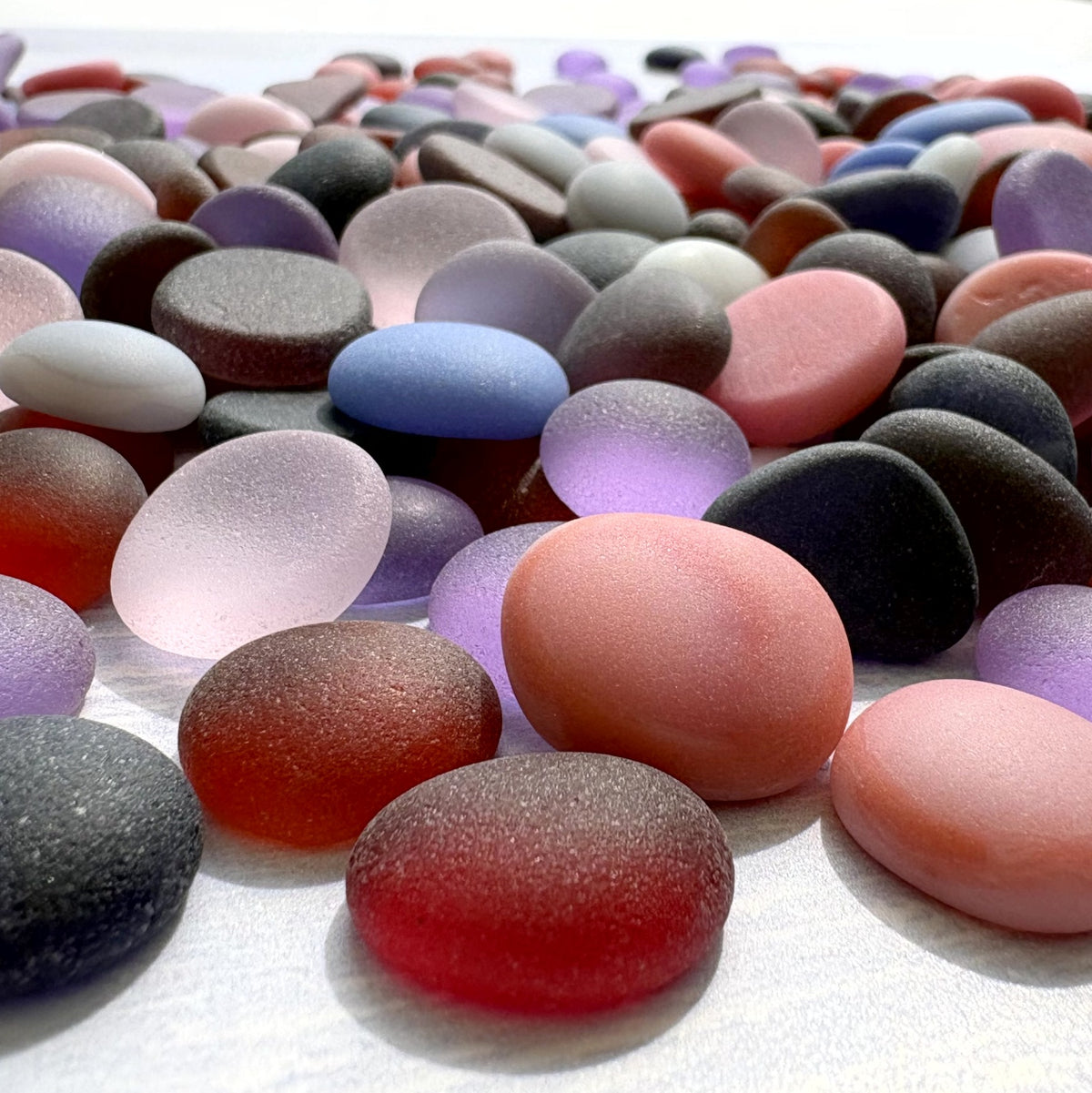 A close-up arrangement of smooth, rounded pebbles and sea glass stones in assorted colors including pink, purple, blue, gray, and white, photographed on a light surface.