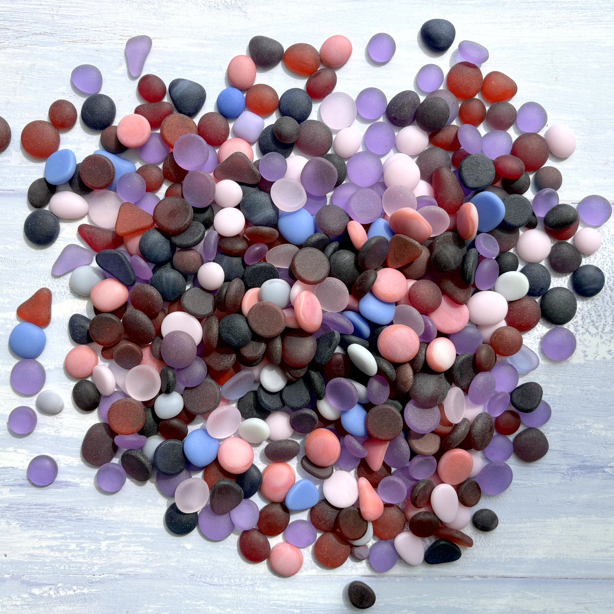 A pile of small, smooth, flat-backed pebbles and faux sea glass cabochons in assorted colours including pink, purple and blue, arranged on a light blue painted wood surface.