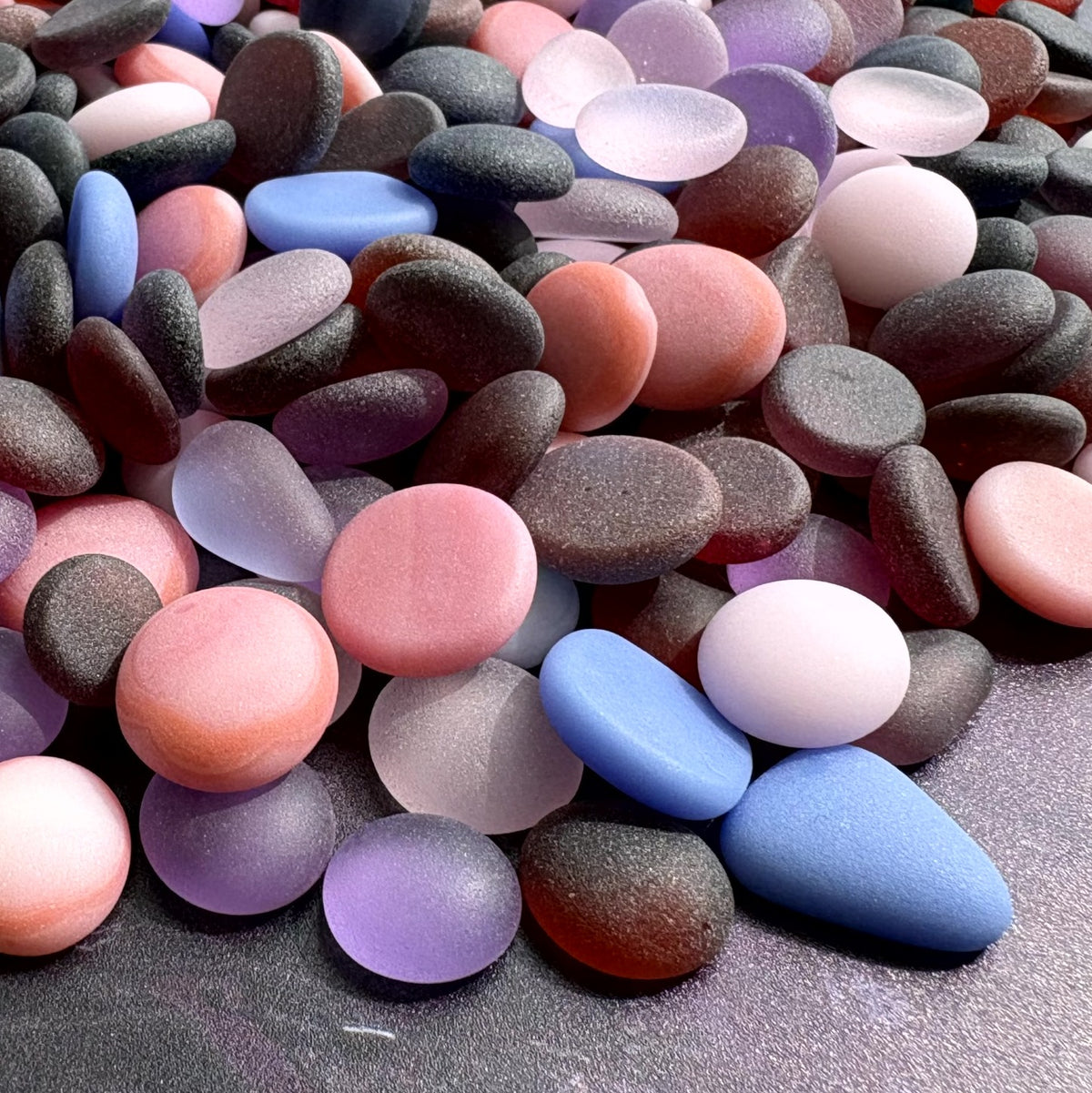 A pile of smooth, rounded decorative stones in assorted colors including pink, and purple tumbled, frosted glass, arranged on a dark textured surface.