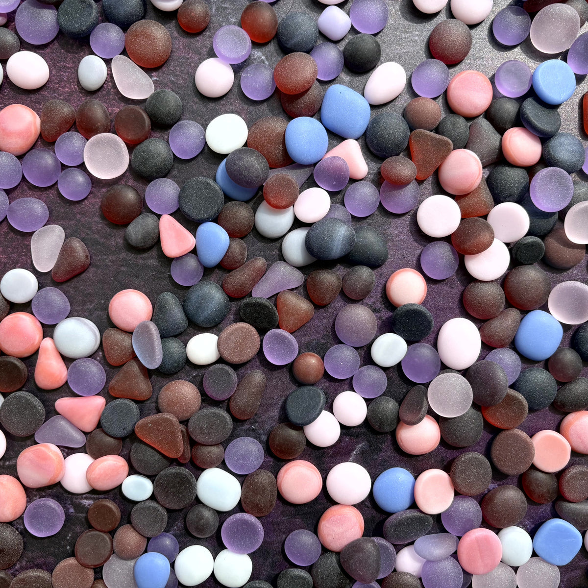 A dense, close-up pile of small, smooth, rounded pebbles and stones in various colors including pink and purple, filling the entire frame.