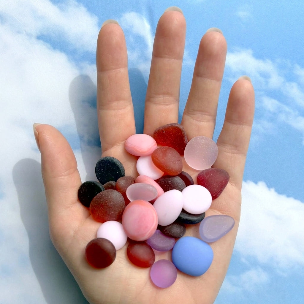 A hand holding a pile of smooth, rounded pebbles and sea glass stones in various colors, including pinks and purples, set against a sky background.