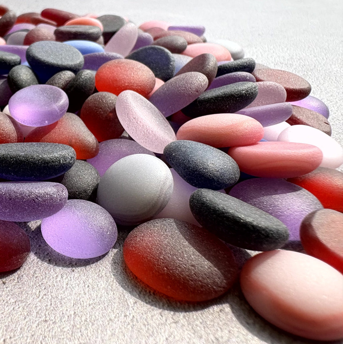 A pile of smooth, polished pebbles and sea glass stones in assorted pastel and neutral colours, including pink, purple, transparent and opaque, arranged on a light textured surface.