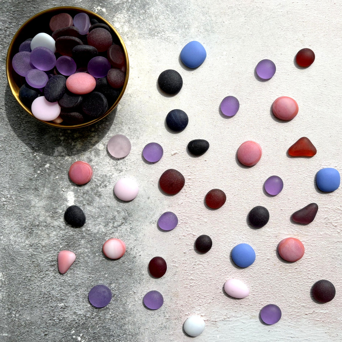 A bowl filled with smooth, polished pebble-shaped frosted glass in assorted colours, with many matching pieces scattered on a light textured surface. The stones come in shades of purple, pink, and blue.