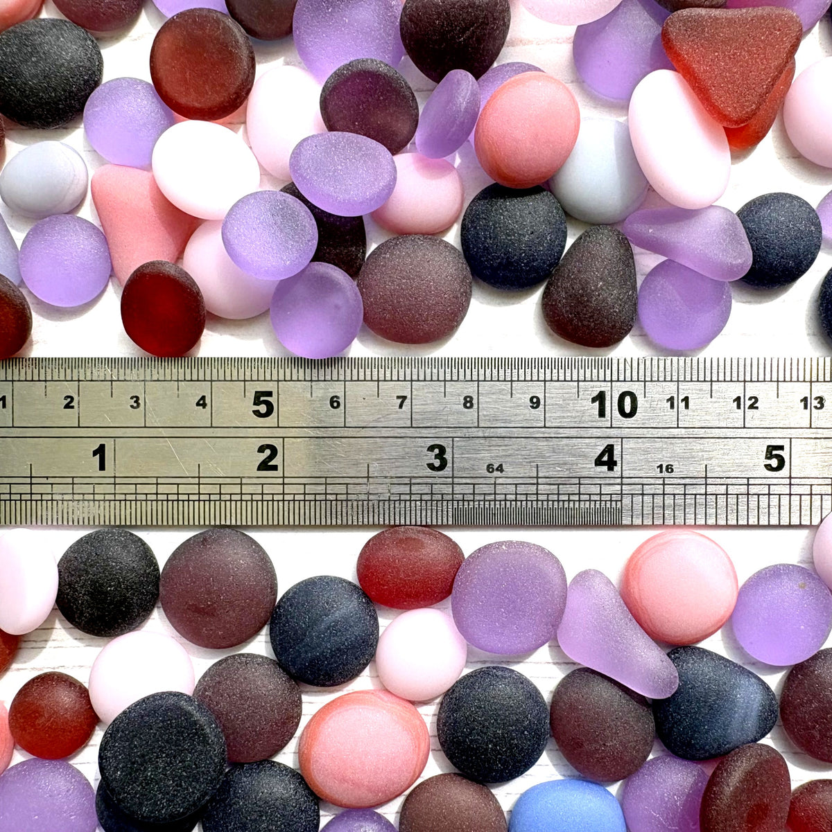 A collection of smooth, rounded beach glass pebbles in various colors including purple, pink, and blue, arranged on a white background with a metal ruler in the middle for scale.