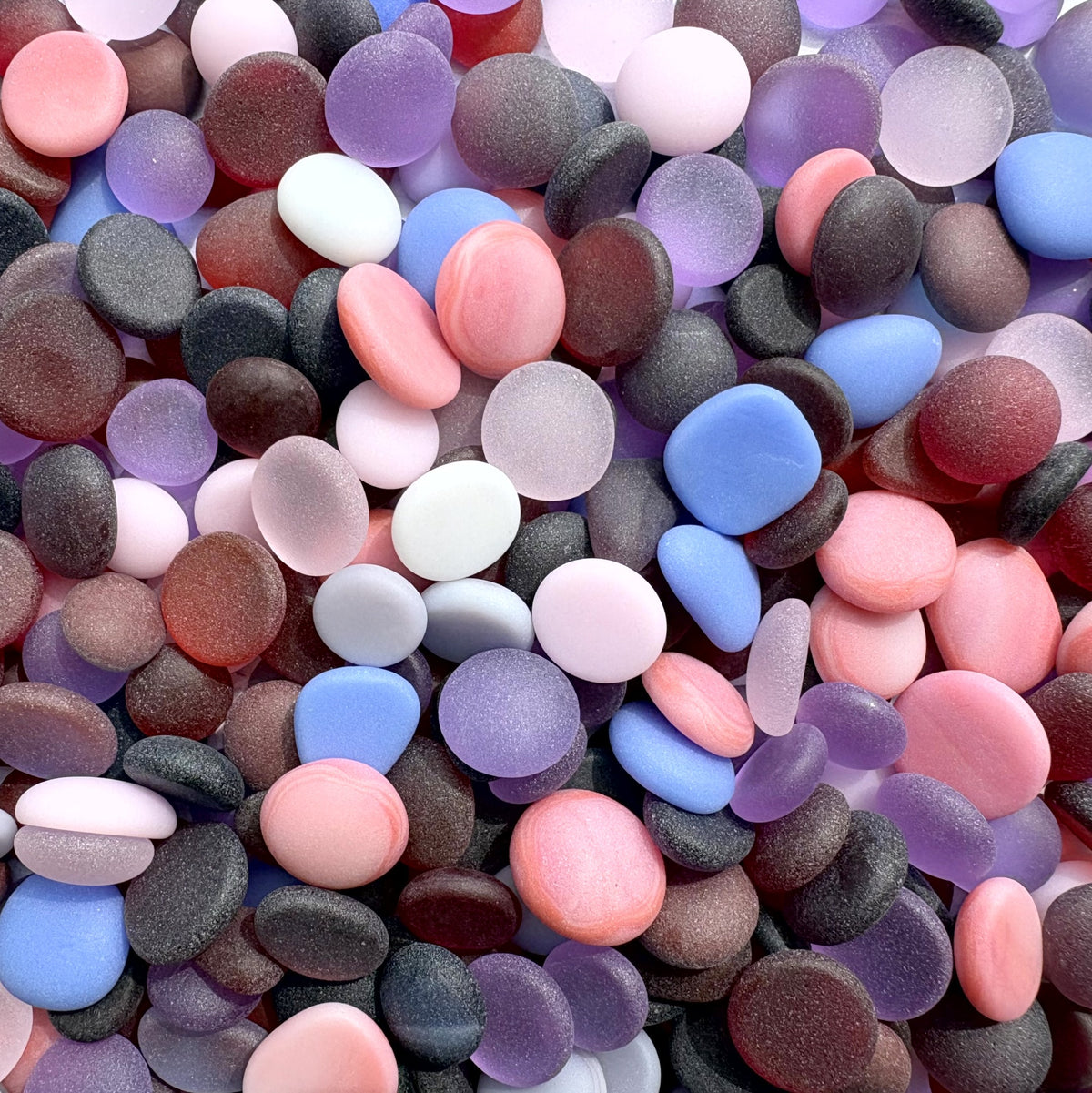 A dense pile of smooth, polished pebble-shaped faux seaglass or decorative stones in assorted pastel and muted colors including pink, blue and purple, lavender, peach, white.