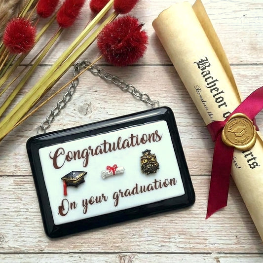 A rectangular handmade glass award picture with a black frame and white centre, hanging from a silver chain. Optional name personalisation. The picture features small 3D graduation-themed additions and the text 'Congratulations On Your Graduation' in brown lettering.