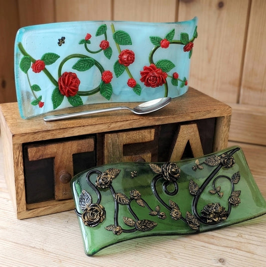 Two decorative glass free standing curved pictures with floral designs, one light blue with red roses and green leaves, and one green with black roses and gold-toned floral accents, displayed on a wooden crate with a spoon on top.