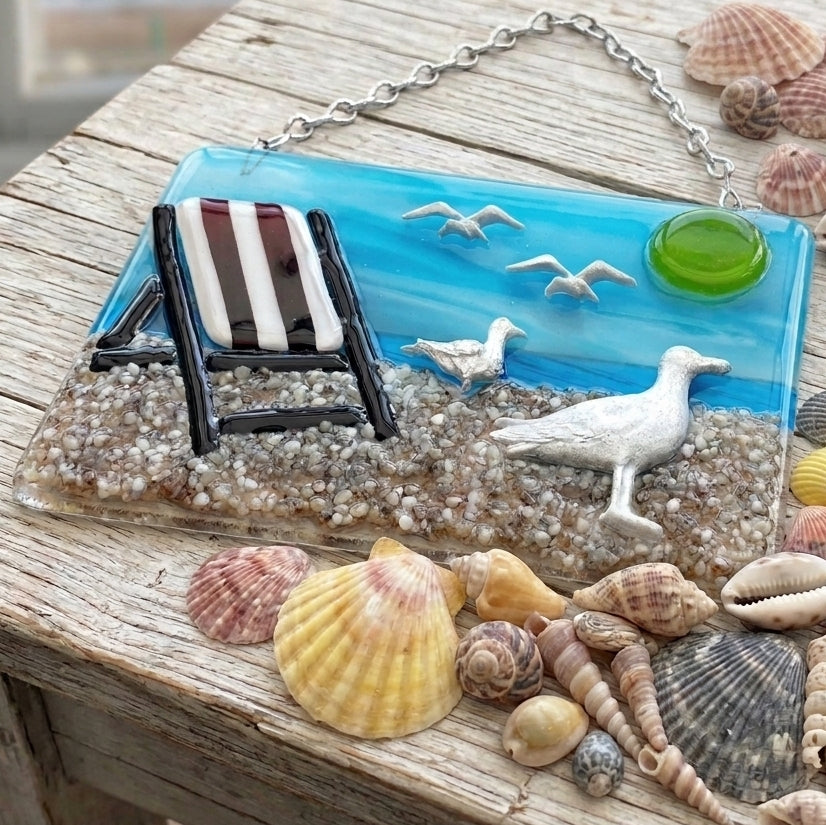 A beach-themed handmade glass plaque or wall hanging with a blue ocean background, seashells, a white seagull, a striped lounge chair, and a silver hanging chain, displayed on a rustic wooden surface with additional shells around it.