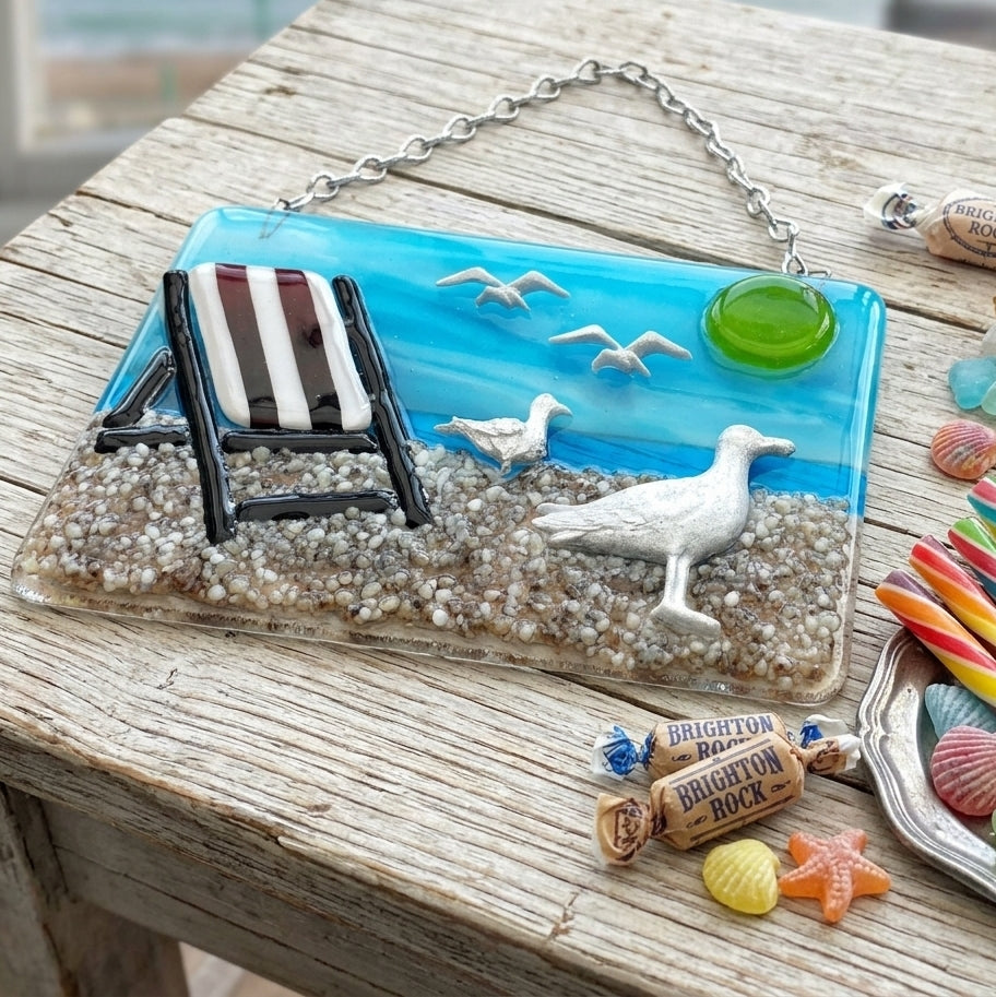 A beach-themed hand made glass suncatcher picture or wall hanging with a silver chain, featuring a blue ocean scene, seagulls, a striped lounge chair, and a textured sandy base. The plaque is displayed on a weathered wooden table with seaside props like Brighton sweets and traditional rock candy.