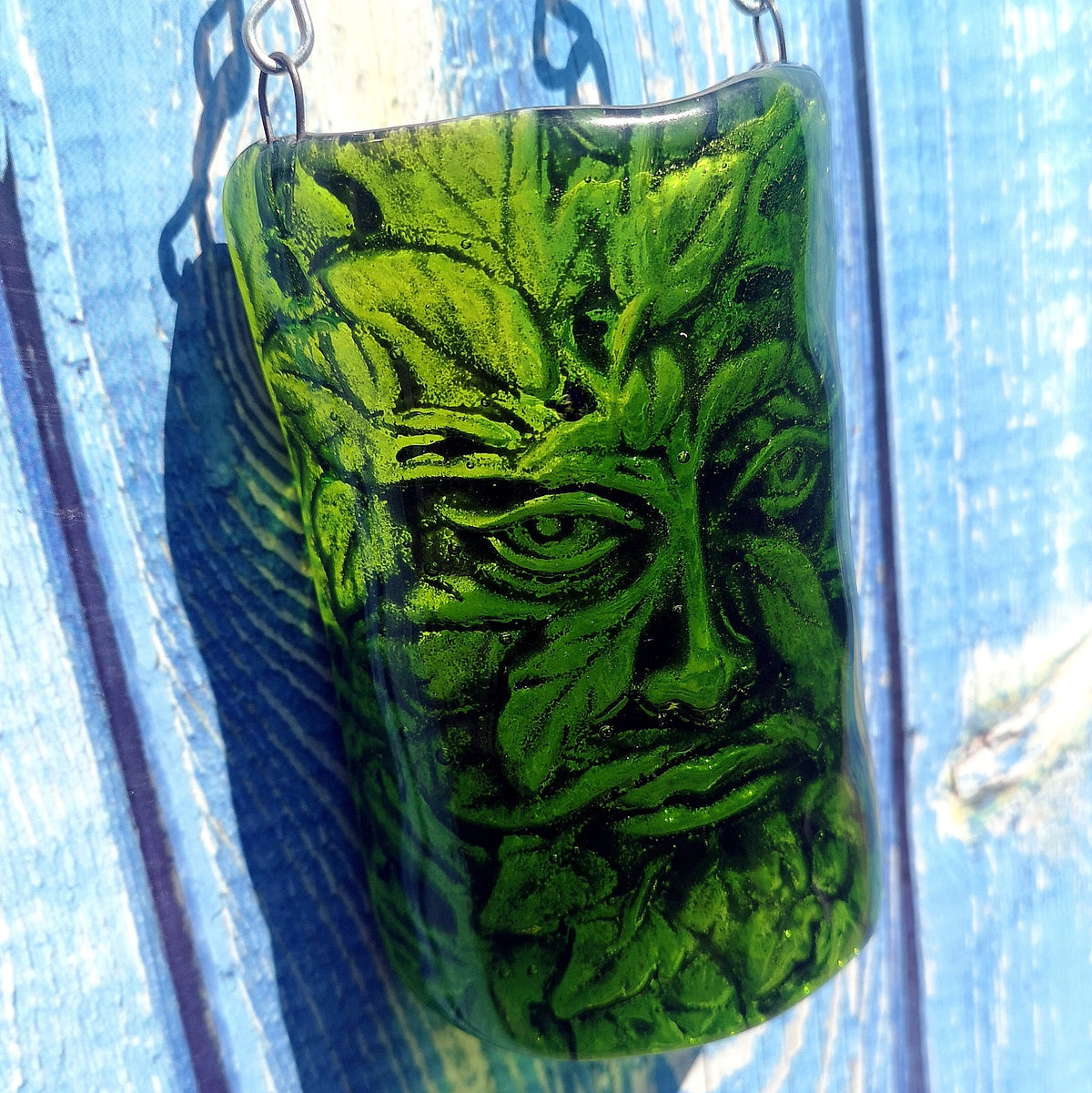 A green glass curved arch picture featuring a carved Leaf Man face, hanging against a weathered blue wooden background. The angle of the picture shows the effect of the raised glass image coming out from the wall.