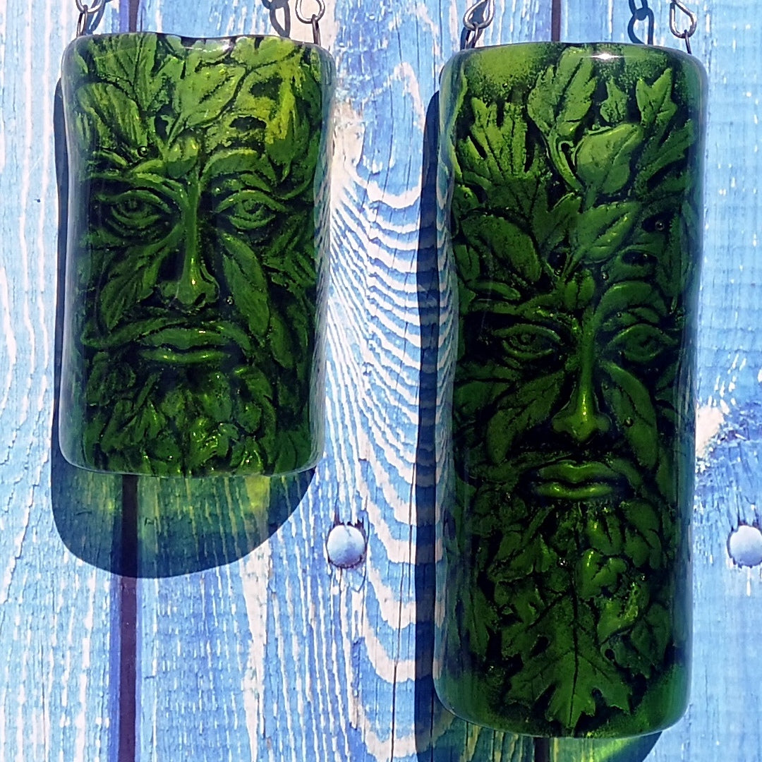 Two green glass hanging wall decorations shaped like a tree trunk with an embossed face made of leaves and a Greenman image, shown hung on chain against a blue wooden plank background.