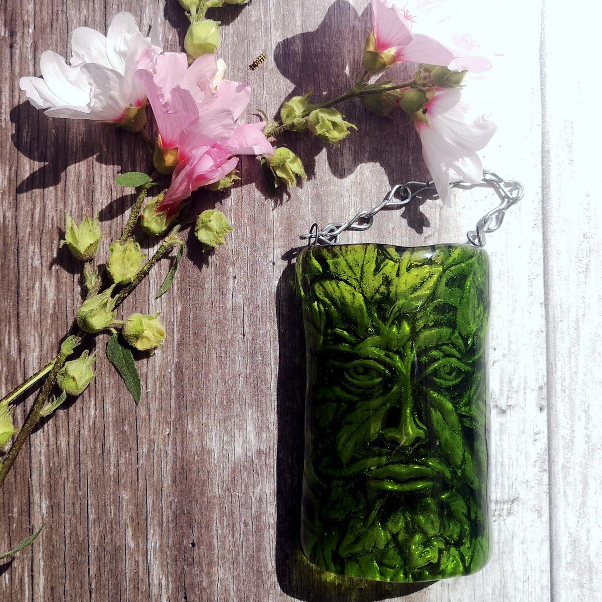 A green glass hanging plaque featuring a carved nature Man Of The Woods face design, displayed on a rustic wooden surface next to pink flowers.