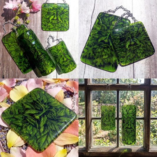A collage showing multiple images of  handmade fused green glass hanging wall plaques with a Greenman face and leaves embedded design, displayed on a wood surface, among pink flowers, and hanging in front of a window.