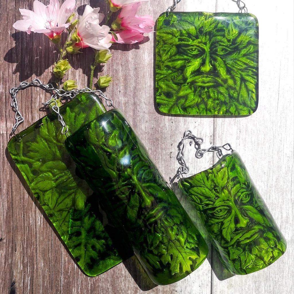 A picture showing the four different options of size and style available for a  green glass hanging picture featuring a Greenman face that has been embedded into the underside of the glass, all displayed on a rustic wood background with pink flowers.