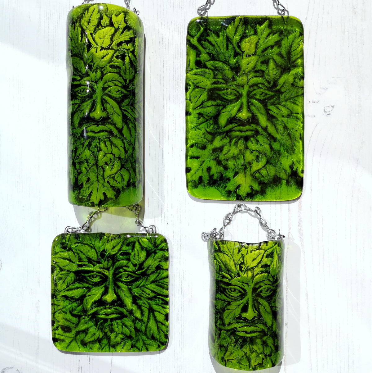 Four green glass hanging ornaments featuring a leafy face nature design, displayed on a white wooden background. Showing the different size and styles available. Can be ordered as a flat hanging plaque, or in a 3D curved arch shape, both in small and large size.