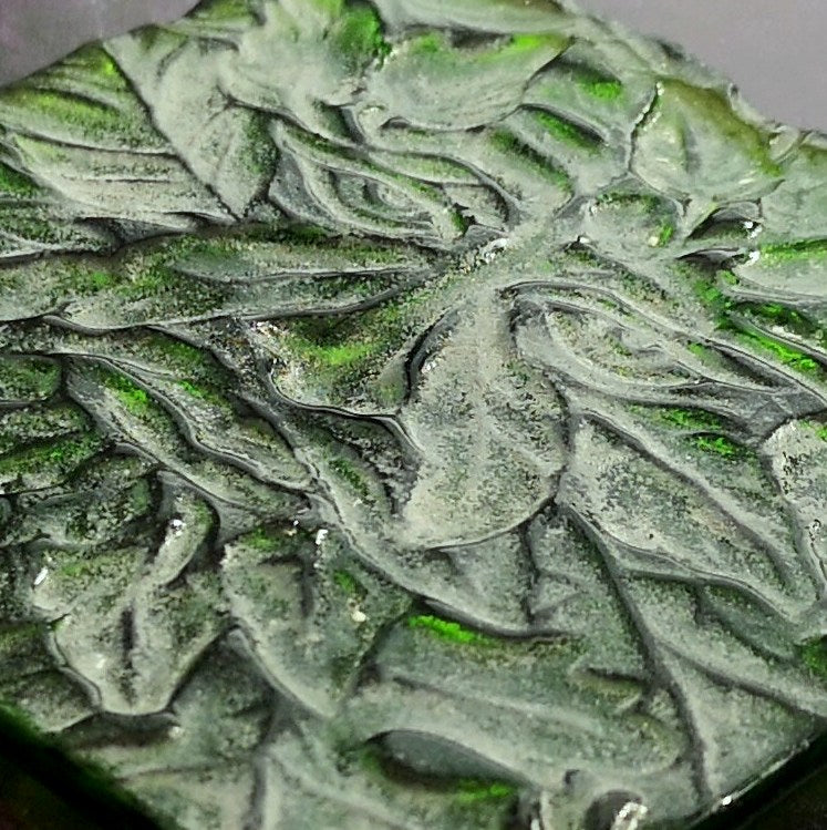 A close-up of the textured underside of an embossed Green Man and leaf pattern set into glass in shades of green and black, with a glossy finish.