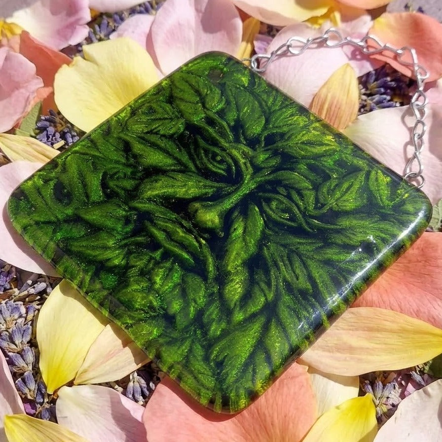 A square green handmade fused glass picture with a raised Man Of The Woods pattern embedded into the glass, and hung with silver chain, resting on pink and yellow flower petals.
