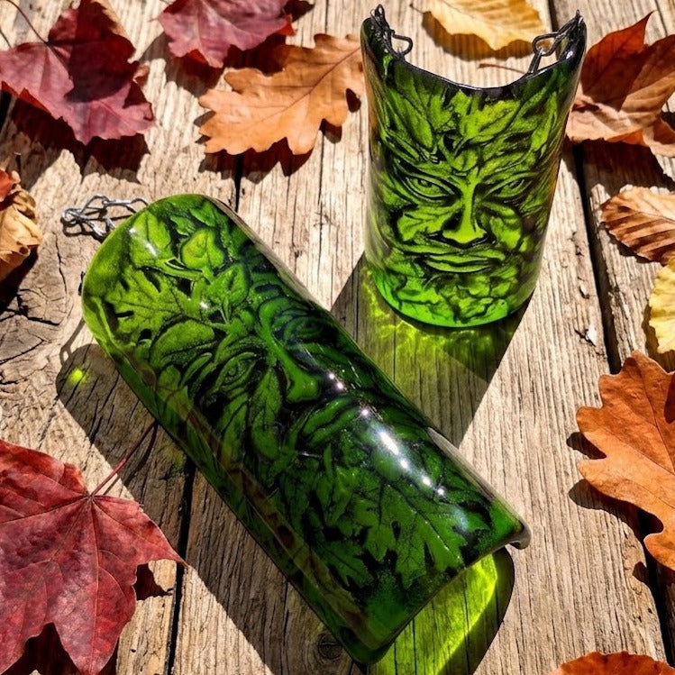 Two green glass three dimensional hanging curved arch pictures with leaf and Greenman face engravings, displayed on a rustic wooden surface surrounded by autumn leaves.