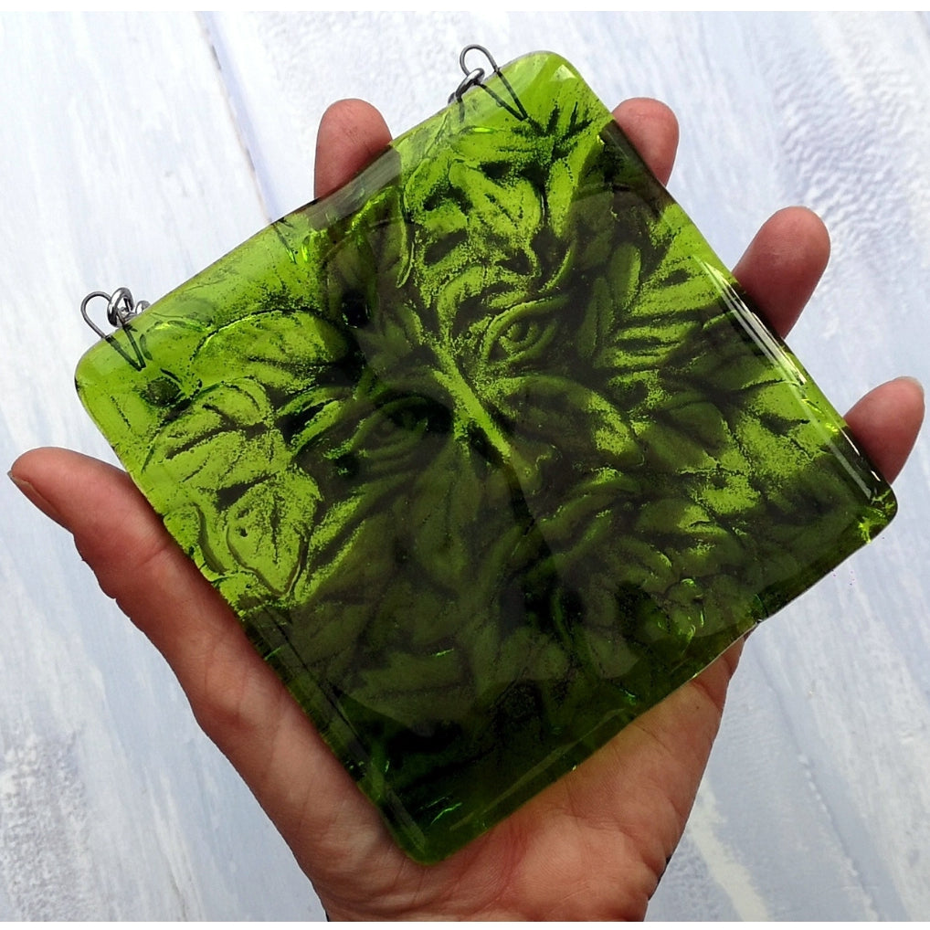 A hand holding a square translucent green glass panel featuring an embossed leaf and Greenman pattern, with two small metal hanging loops at the top corners, set against a light grey painted wood background.