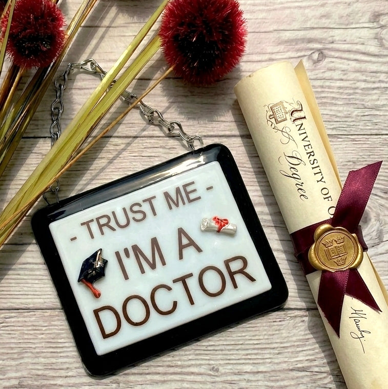 A rectangular handmade glass award picture with a black frame and white centre, hanging from a silver chain. Optional name personalisation. The picture features small 3D graduation-themed additions and the text 'Trust Me - I'm A Doctor' in brown lettering.