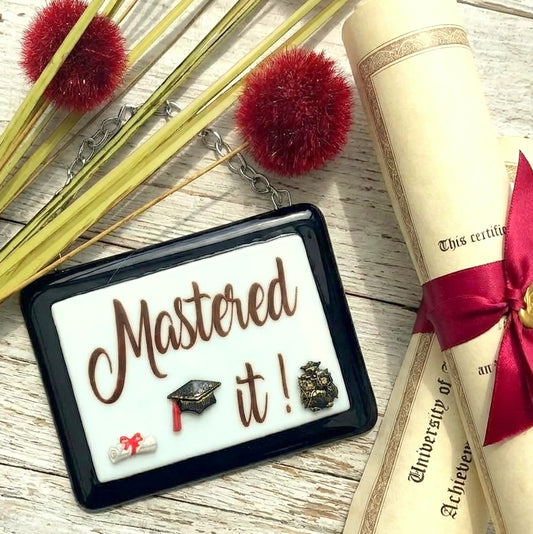A rectangular handmade glass award picture with a black frame and white centre, hanging from a silver chain. Optional name personalisation. The picture features small 3D graduation-themed additions and the text 'Mastered It!' in brown lettering.