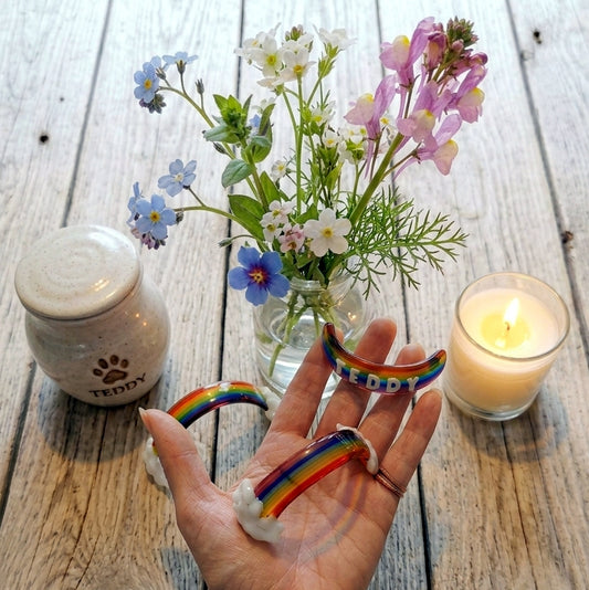 Three small handmade glass rainbow bridge pet memorials with clouds and personalised with optional 3D names,  displayed on a person's open hand, surrounded by flowers in a glass vase, a lit candle, and a ceramic pet ashes jar on a rustic wooden surface.