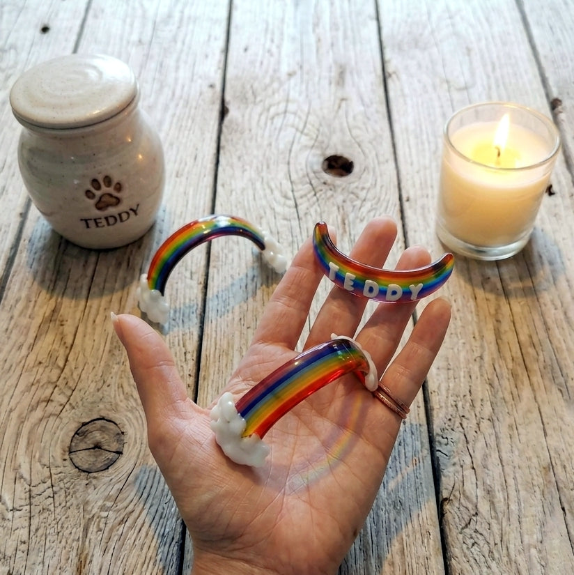 A hand holds three small handmade glass rainbow bridge pet memorials with clouds and personalised with optional 3D names, set on a rustic wooden surface with a candle and a ceramic pet ashes jar in the background.