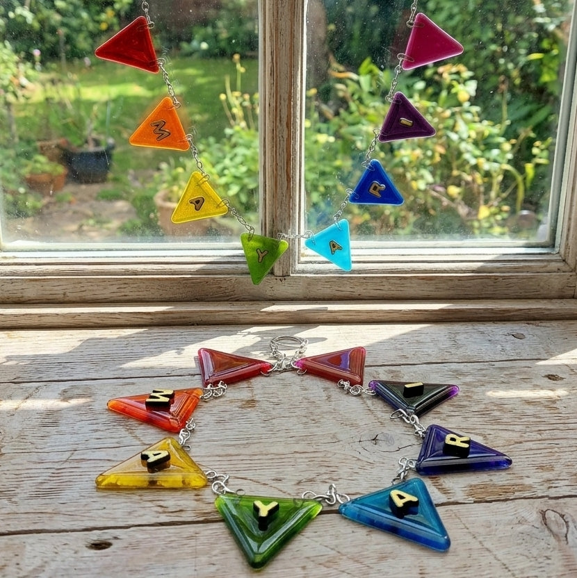 A colourful handmade glass sunburst personalised name bunting and matching suncatcher garland displayed on a rustic wooden windowsill with a garden background visible through the window.