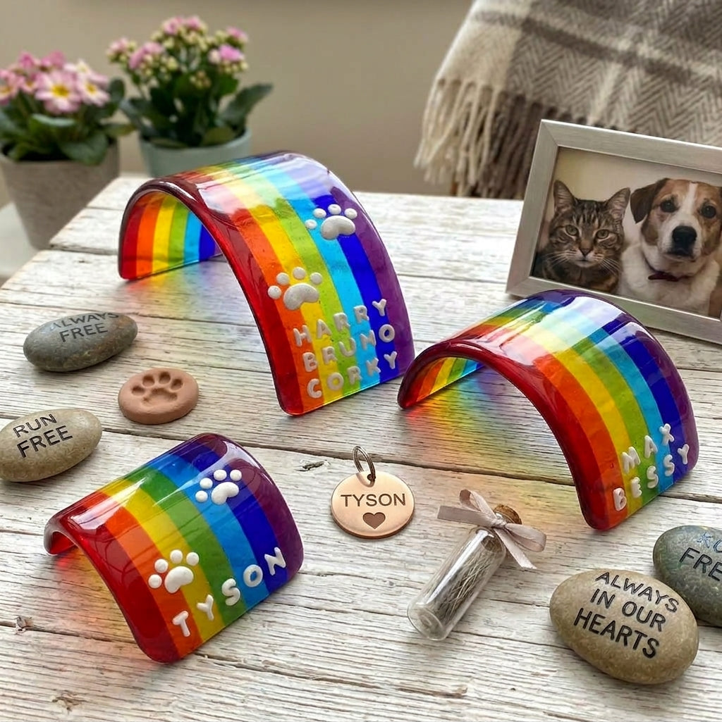Three colourful glass curved rainbow bridge pet memorial keepsake items with rainbow stripes and white 3D paw prints and personalised multiple names, displayed on a rustic wooden surface with engraved stones, small flowers, a blanket, and a pet photo frame in the background.