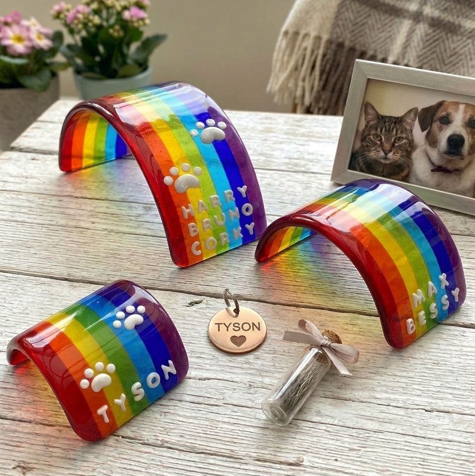 Three colourful glass curved rainbow bridge pet memorial keepsake items with rainbow stripes and white 3D paw prints and personalised multiple names, displayed on a rustic wooden surface with  small flowers, a blanket, and a pet photo frame in the background.