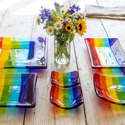 A set of handmade fused glass rainbow-striped tableware arranged on a light wood table, including a rectangular plate, two variations of small bowls, and a matching medium sized bowl, plus large and extra large sized square dishes. With a glass vase of mixed flowers in the center.