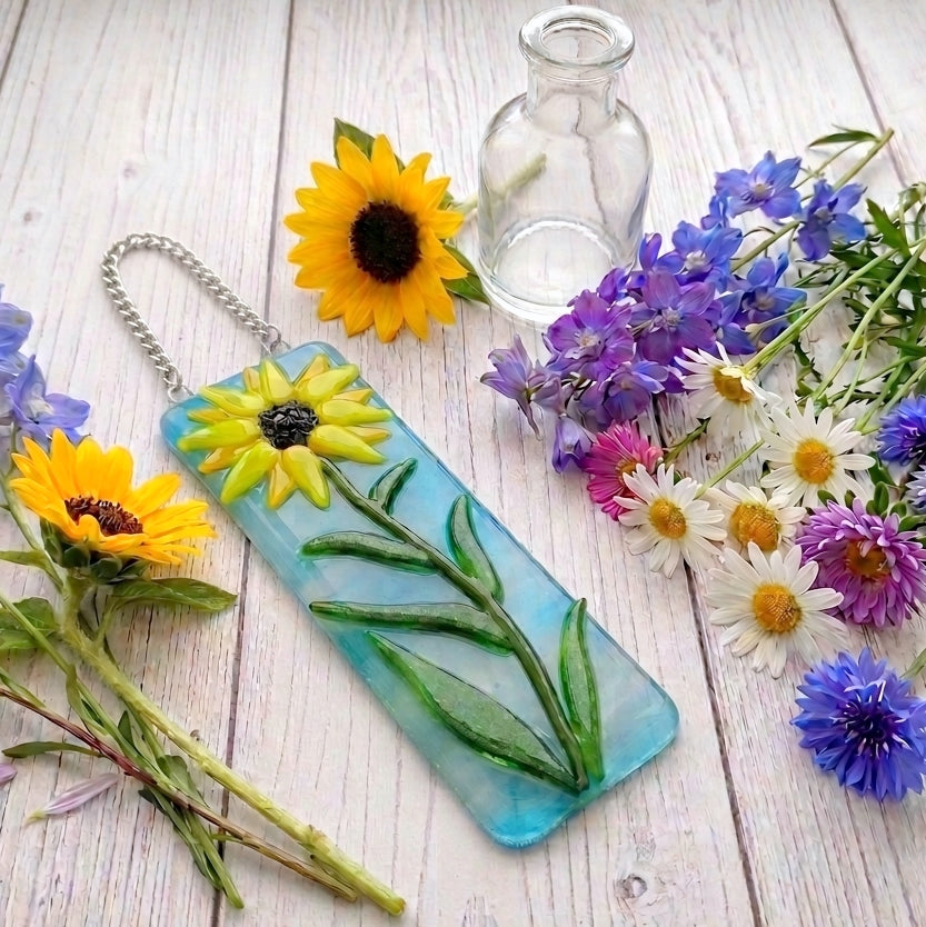A rectangular glass suncatcher keepsake tag with a yellow sunflower design and green stem and leaf details, attached to a silver chain. The tag is displayed on a white wooden surface surrounded by real sunflowers, purple and white wildflowers, and a small clear glass bottle.