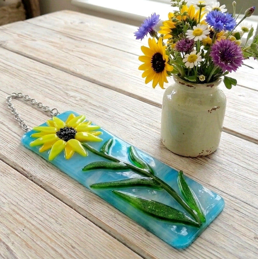 A rectangular turquoise glass plaque featuring a raised yellow sunflower with a dark centre and green stem, attached to a silver chain. The plaque is displayed on a light wood table top next to a ceramic vase filled with yellow, purple, and white flowers.