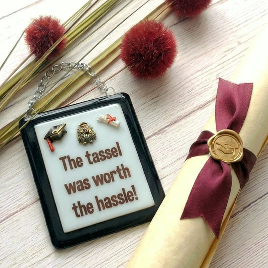 A rectangular handmade glass award picture with a black frame and white centre, hanging from a silver chain. Optional name personalisation. The picture features small 3D graduation-themed additions and the text 'The tassel was worth the hassle!' in brown lettering.