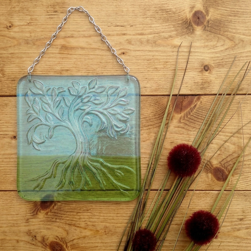 A square glass hanging plaque featuring an embossed tree of life with a blue sky and green grass design, suspended by a silver chain on a wooden surface next to decorative reeds. Handmade fused glass Tree Of Life hanging picture. Wall plaque suncatcher, nature. Iridised glass, sacred tree light catcher.