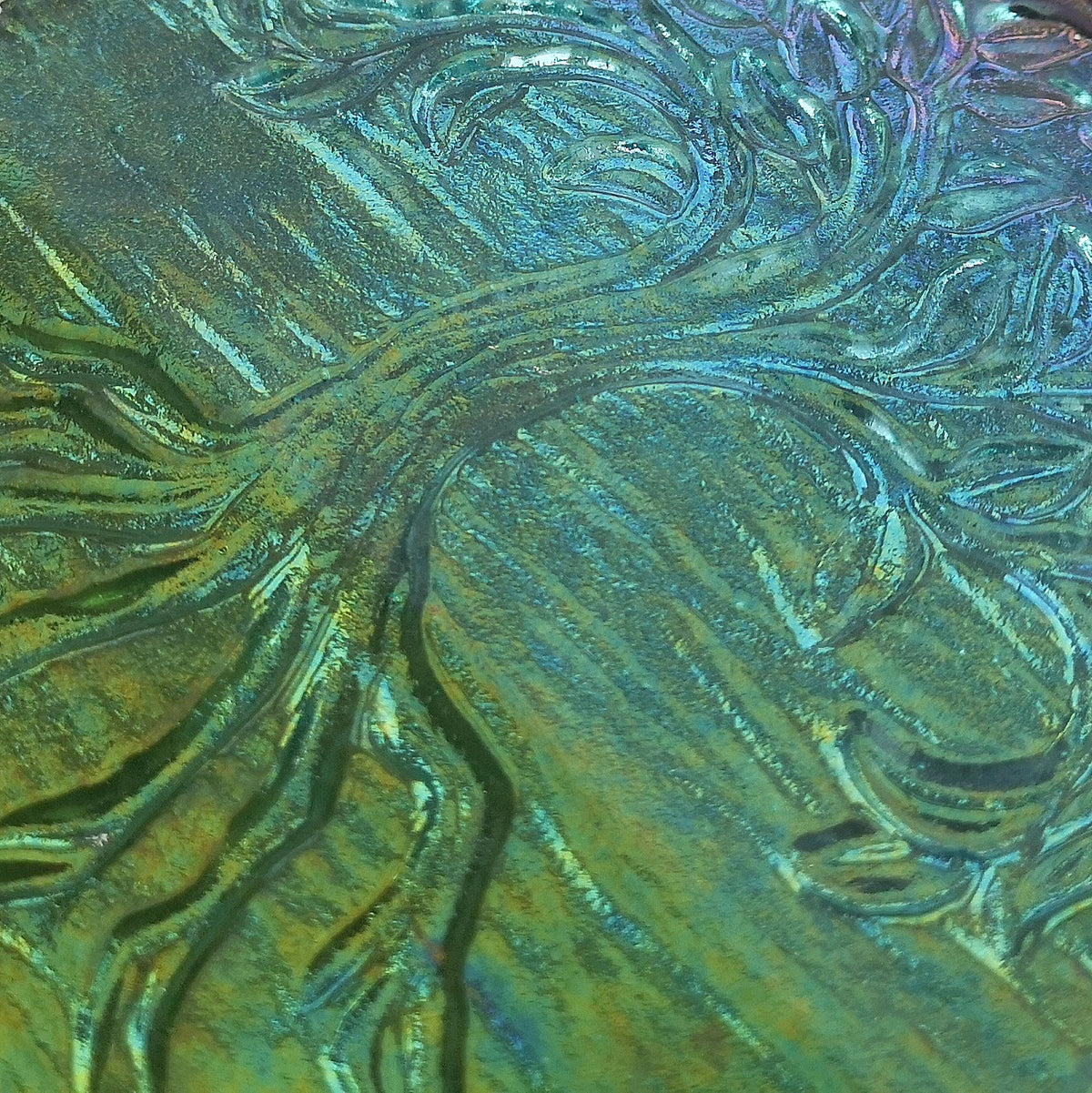 A close-up of an embossed tree design with swirling branches and roots, featuring an iridescent finish that shifts between green, blue, and gold tones. This is the reverse side of a handmade fused glass Tree Of Life hanging picture. Wall plaque suncatcher, nature. Iridised glass, sacred tree light catcher.