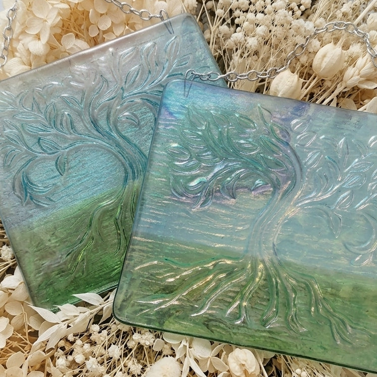 Two square glass wall art panels featuring an embossed tree of life design, displayed on a bed of dried cream flowers. The glass has a gradient colour shift from light blue at the top to green at the bottom, with a silver hanging chain visible at the top.