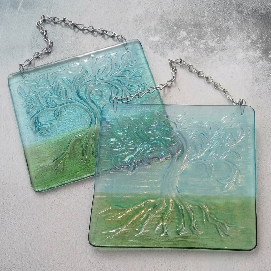 Two rectangular glass hanging plaques with a raised tree of life design, featuring a blue-to-green gradient and silver hanging chains, displayed on a textured light grey surface.
