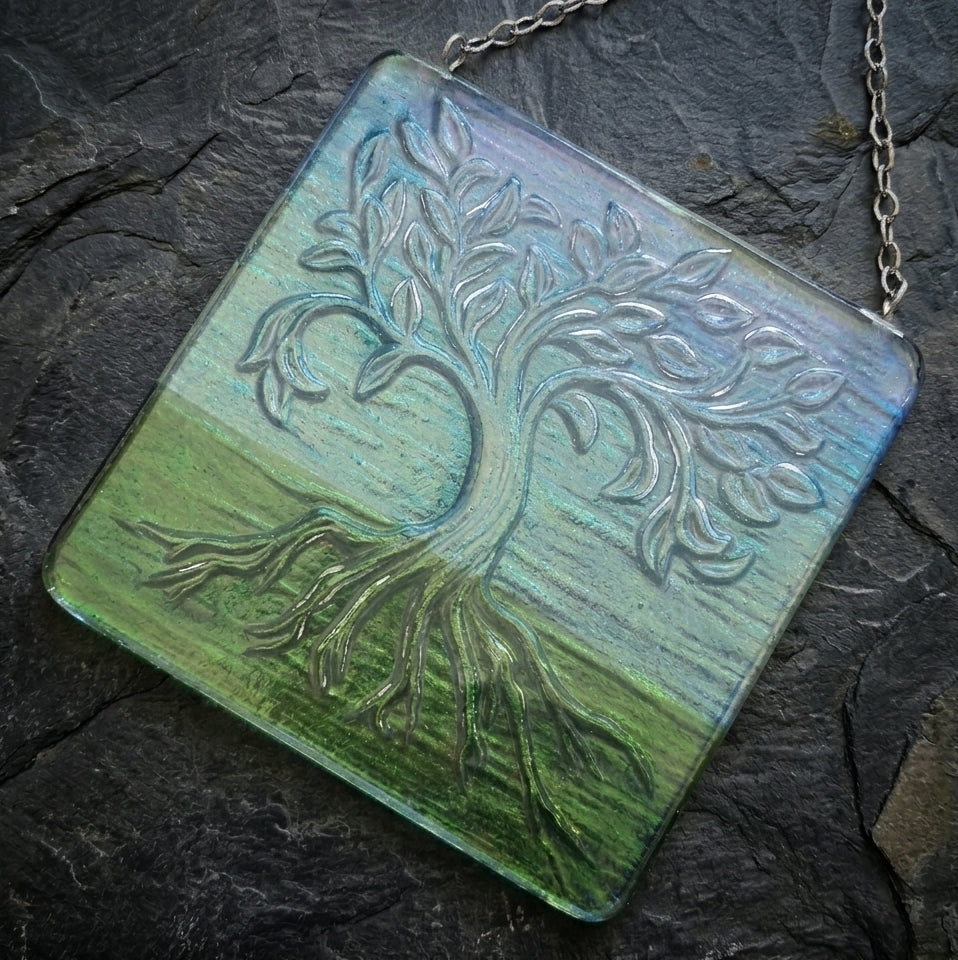 A square glass picture featuring an embossed tree of life design over a textured background that transitions from light blue at the top to green at the bottom, hanging from a silver chain on a dark slate surface.