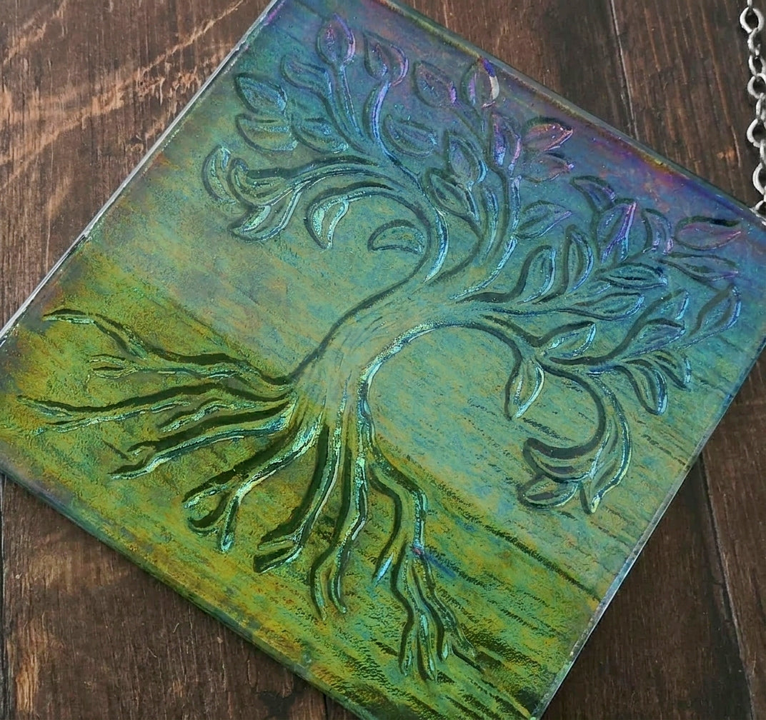A square glass decorative hanging wall plaque featuring an embossed tree of life design, resting on a dark wood surface. The glass has a vibrant green and blue iridescent finish with subtle purple and orange shifts.