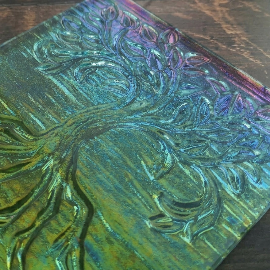 A close-up of a square decorative plaque or tile featuring an embossed tree design with an iridescent finish shifting between green, blue, purple, and gold, placed on a dark wood surface.
