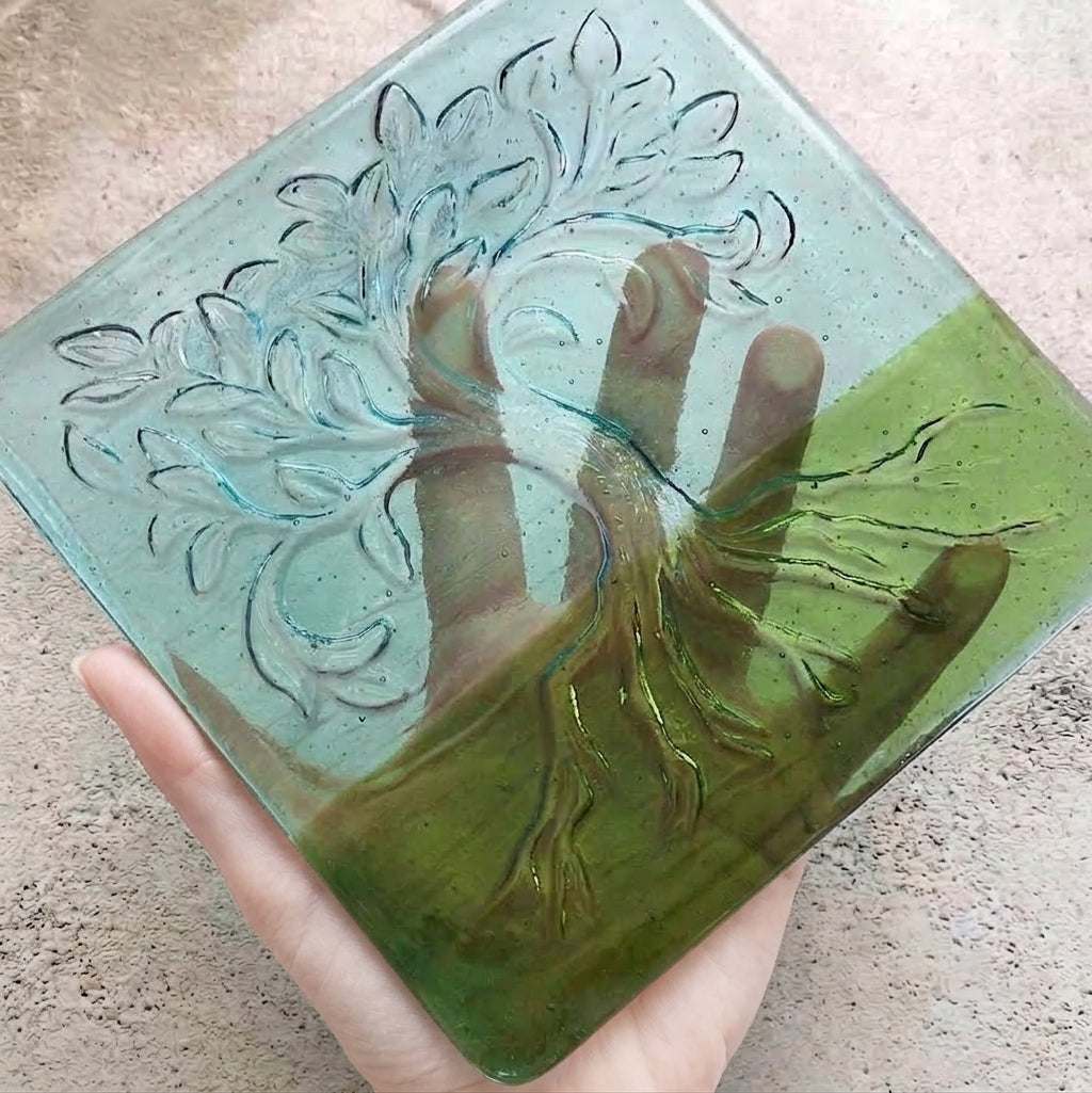 A hand holds a square glass art piece featuring an embossed tree of life design. The glass has a light blue-green top half and a darker green bottom half, representing the sky and grass.